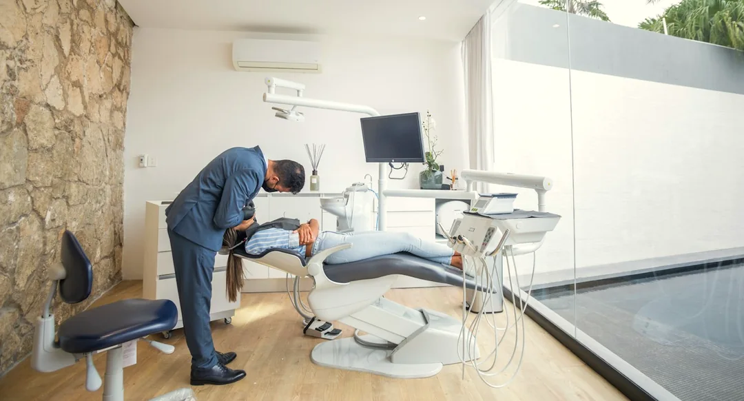 Dentist examining a patient in a sunlit treatment room with natural stone walls, wood floors, and garden views